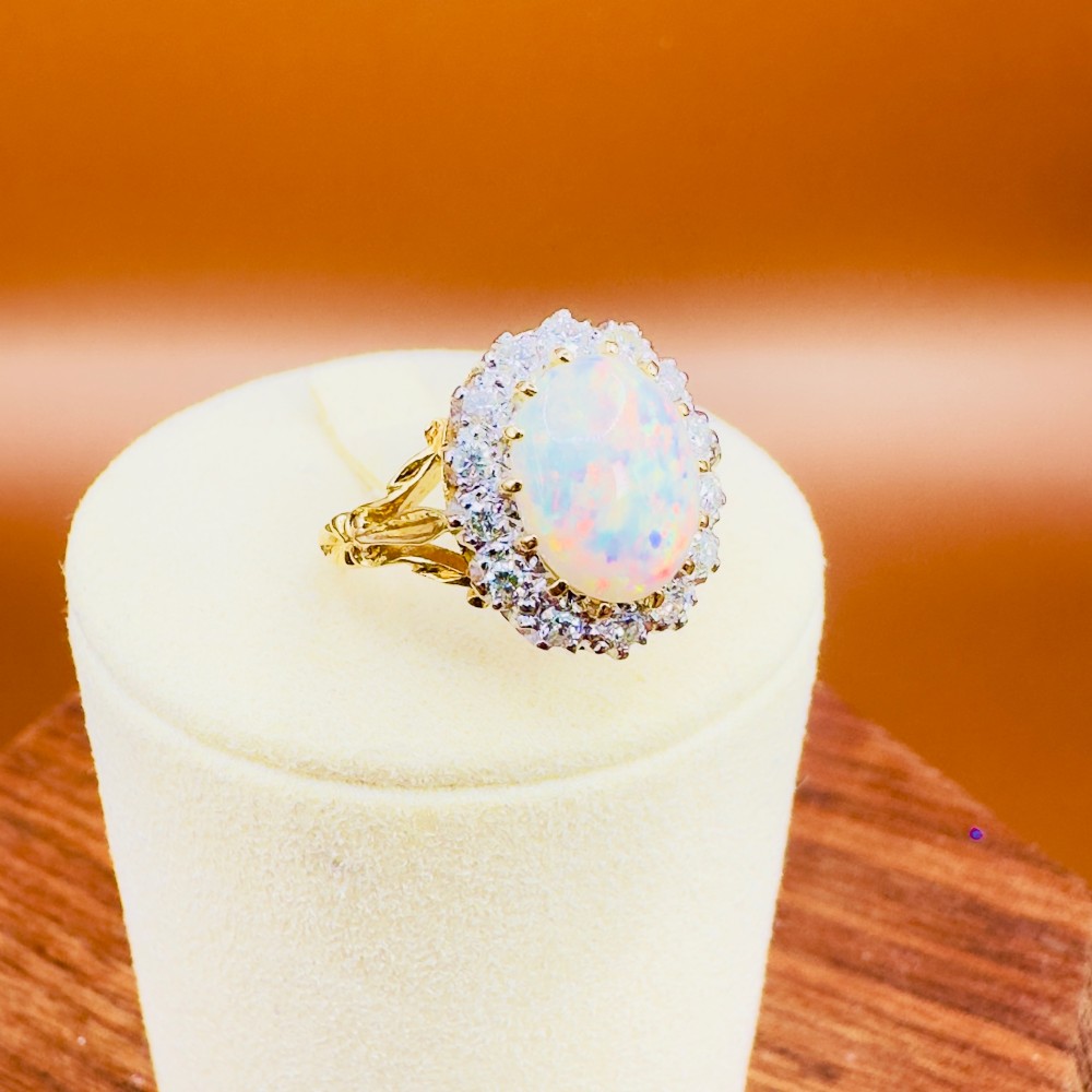 An 18ct yellow gold ring featuring a central oval cabochon opal surrounded by a halo cluster of small round brilliant cut diamonds. View X