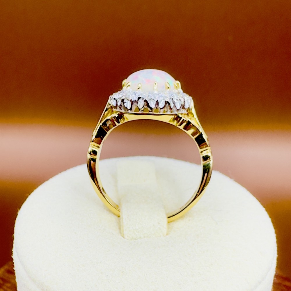 An 18ct yellow gold ring featuring a central oval cabochon opal surrounded by a halo cluster of small round brilliant cut diamonds. View XI