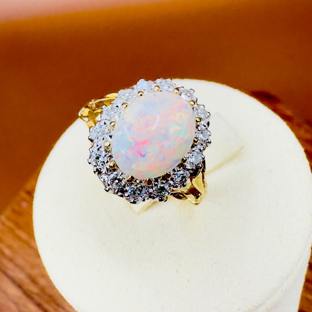 An 18ct yellow gold ring featuring a central oval cabochon opal surrounded by a halo cluster of small round brilliant cut diamonds. View XII
