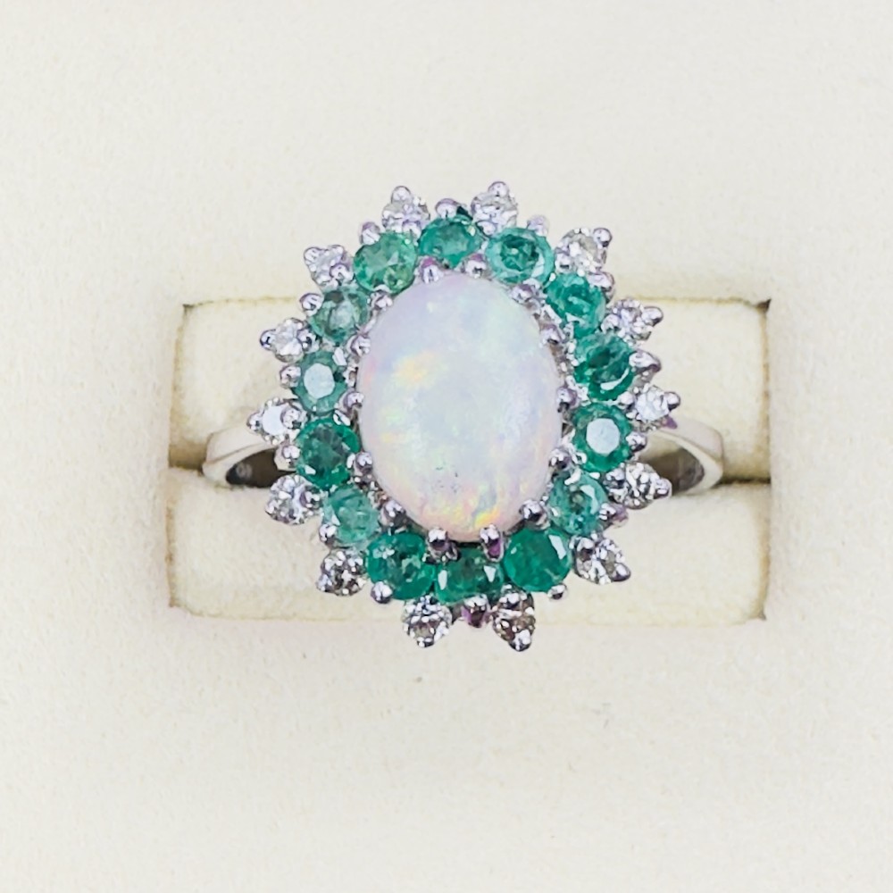 An 18ct white gold cluster cocktail ring set with a central oval cabochon opal framed by a halo of round emeralds and accent diamonds. View II