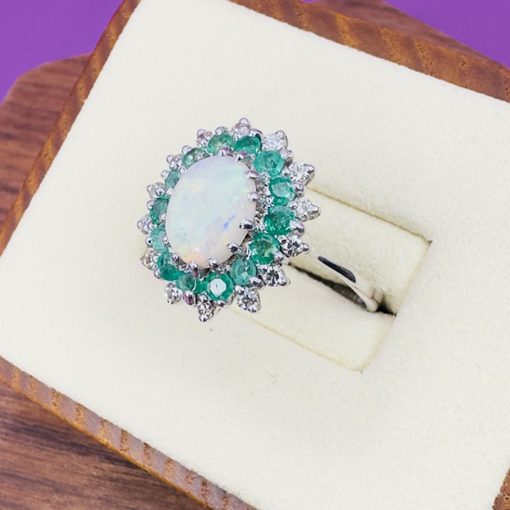 An 18ct white gold cluster cocktail ring set with a central oval cabochon opal framed by a halo of round emeralds and accent diamonds. View III