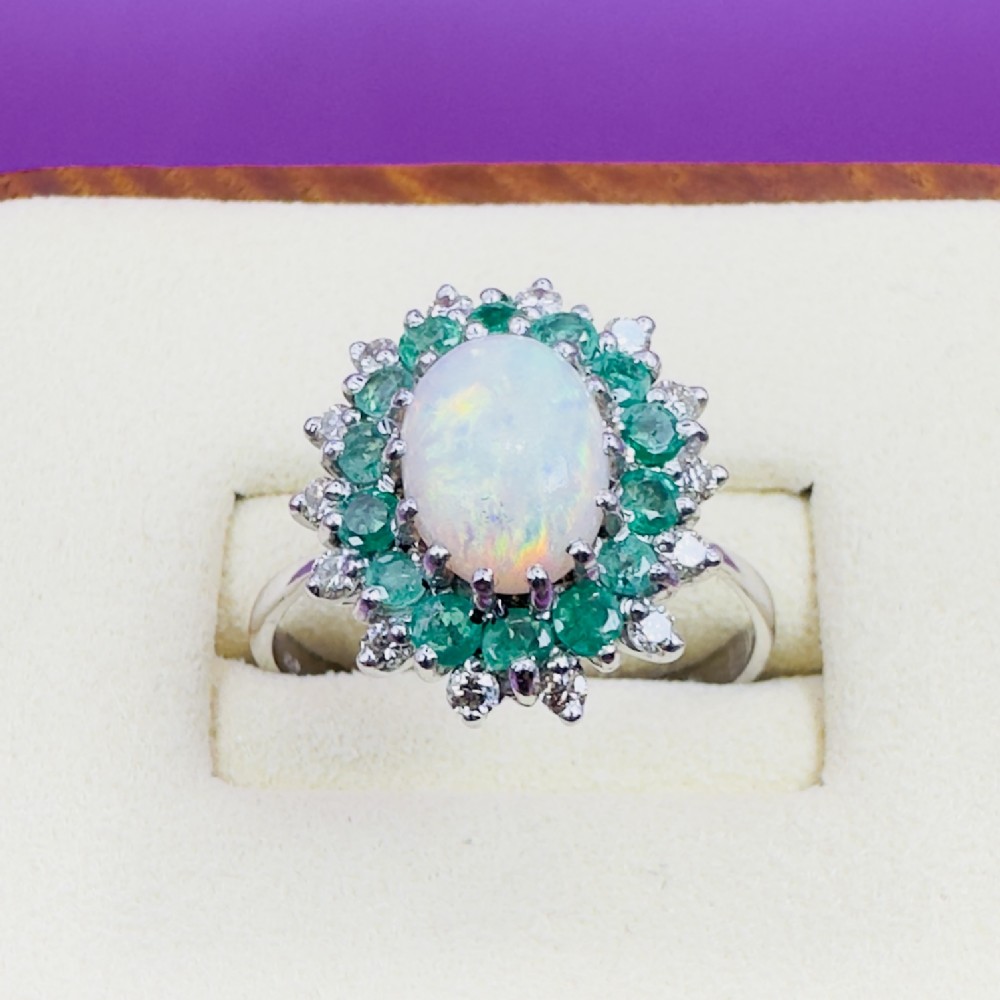 An 18ct white gold cluster cocktail ring set with a central oval cabochon opal framed by a halo of round emeralds and accent diamonds. View V