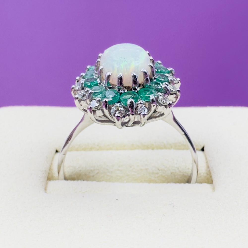 An 18ct white gold cluster cocktail ring set with a central oval cabochon opal framed by a halo of round emeralds and accent diamonds. View VI