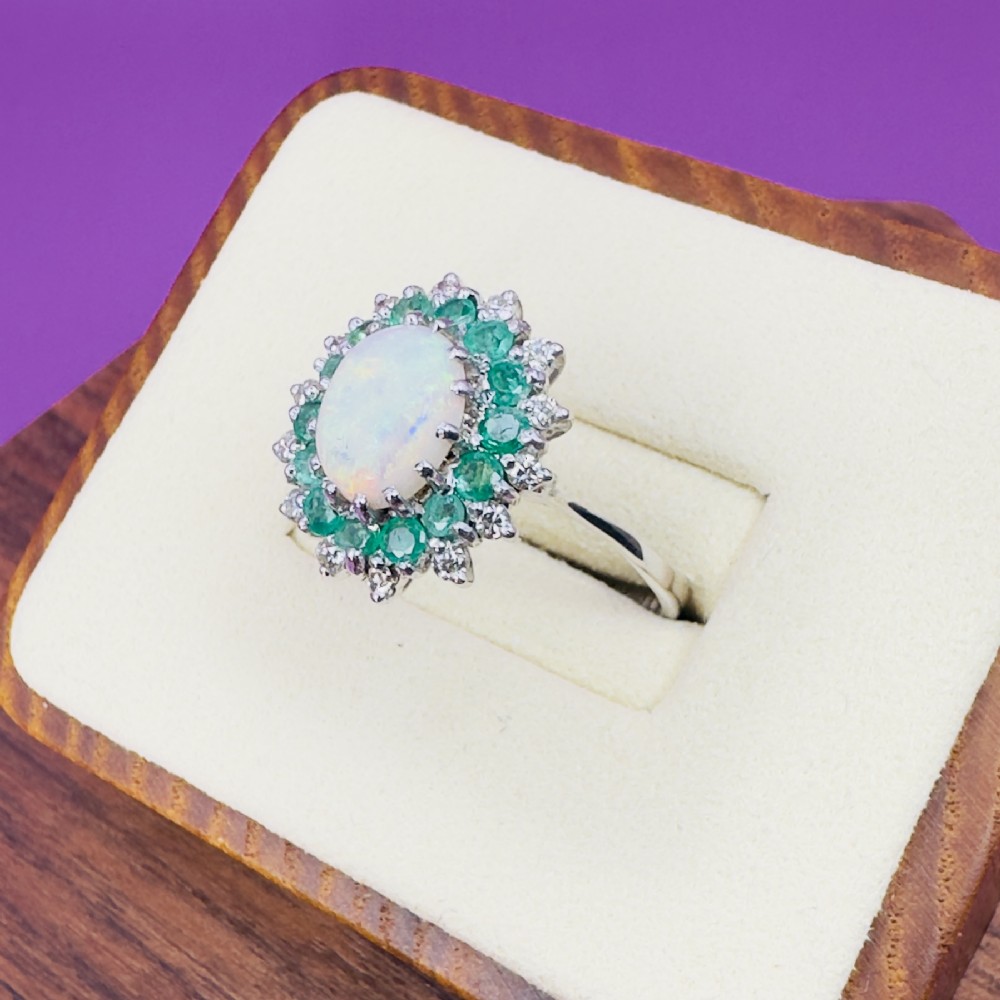 An 18ct white gold cluster cocktail ring set with a central oval cabochon opal framed by a halo of round emeralds and accent diamonds. View VII