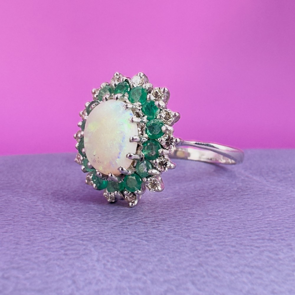 An 18ct white gold cluster cocktail ring set with a central oval cabochon opal framed by a halo of round emeralds and accent diamonds. View VIII