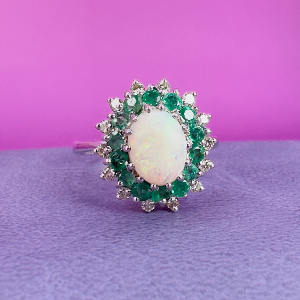 An 18ct white gold cluster cocktail ring set with a central oval cabochon opal framed by a halo of round emeralds and accent diamonds. View IX