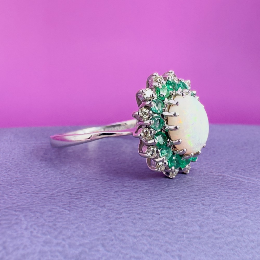 An 18ct white gold cluster cocktail ring set with a central oval cabochon opal framed by a halo of round emeralds and accent diamonds. View X