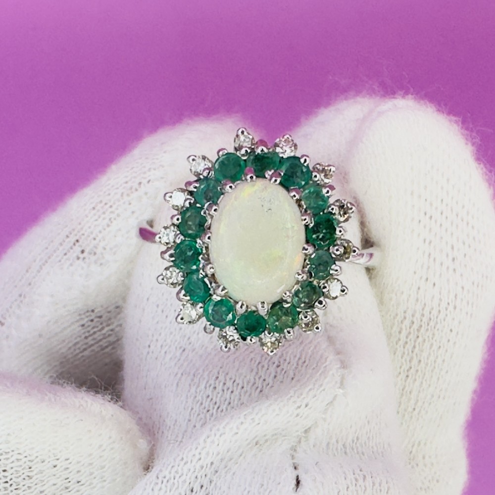 An 18ct white gold cluster cocktail ring set with a central oval cabochon opal framed by a halo of round emeralds and accent diamonds. View XI