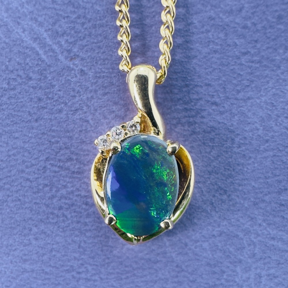 An 18ct yellow gold necklace featuring an oval cabochon black opal displaying green and blue play-of-colour, framed in a contoured gold setting with small diamond accents. View V