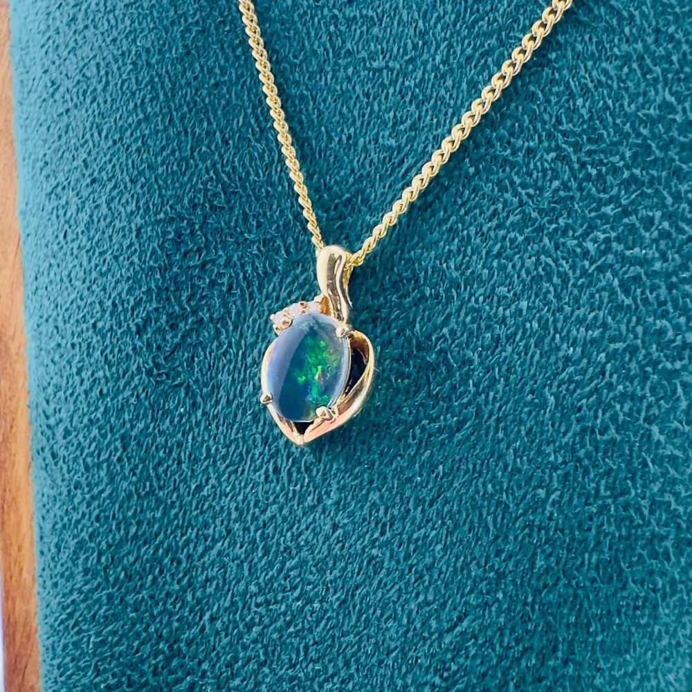 An 18ct yellow gold necklace featuring an oval cabochon black opal displaying green and blue play-of-colour, framed in a contoured gold setting with small diamond accents. View IX
