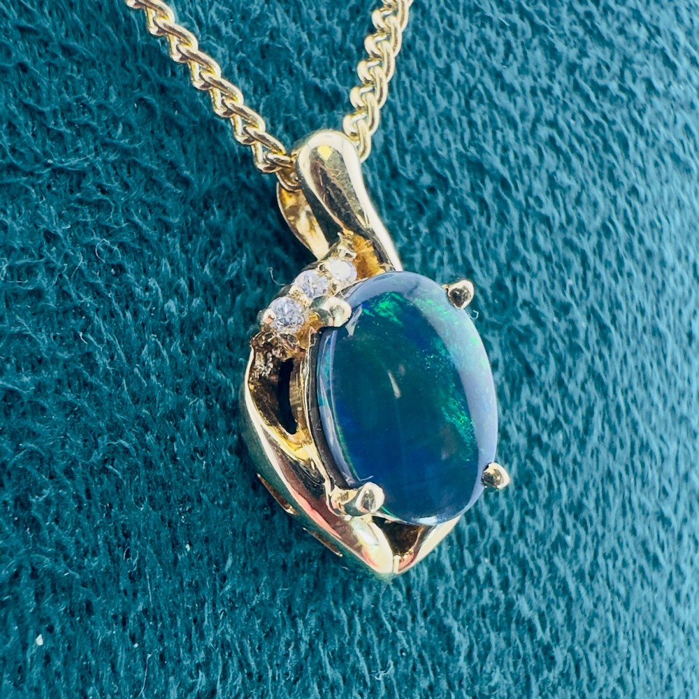 An 18ct yellow gold necklace featuring an oval cabochon black opal displaying green and blue play-of-colour, framed in a contoured gold setting with small diamond accents. View XII