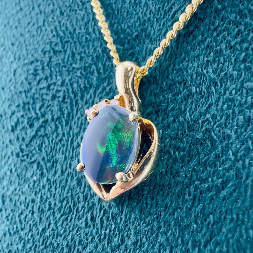 An 18ct yellow gold necklace featuring an oval cabochon black opal displaying green and blue play-of-colour, framed in a contoured gold setting with small diamond accents. View XIII