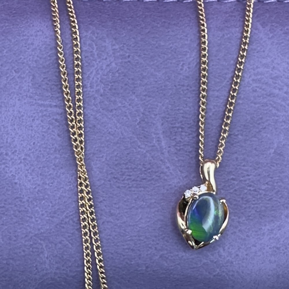 An 18ct yellow gold necklace featuring an oval cabochon black opal displaying green and blue play-of-colour, framed in a contoured gold setting with small diamond accents. View XV