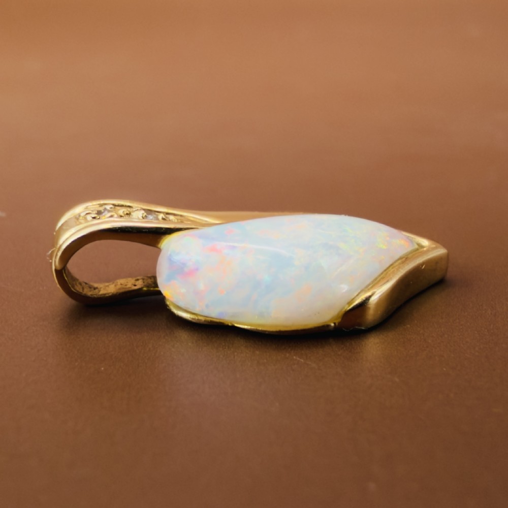 An 18ct yellow gold freeform pendant displaying an iridescent white opal with green and pink flash, set alongside two accent diamonds on the bail. View VIII