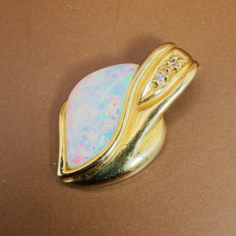 An 18ct yellow gold freeform pendant displaying an iridescent white opal with green and pink flash, set alongside two accent diamonds on the bail. View X