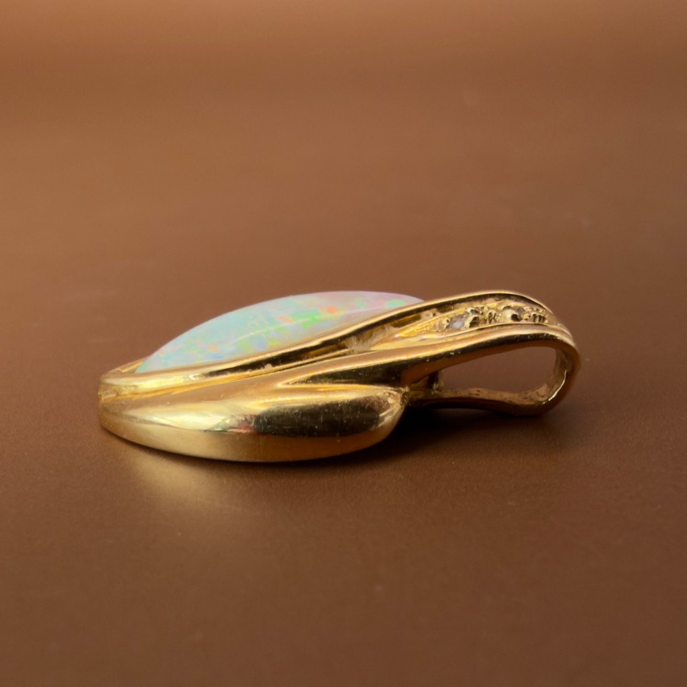 An 18ct yellow gold freeform pendant displaying an iridescent white opal with green and pink flash, set alongside two accent diamonds on the bail. View XI