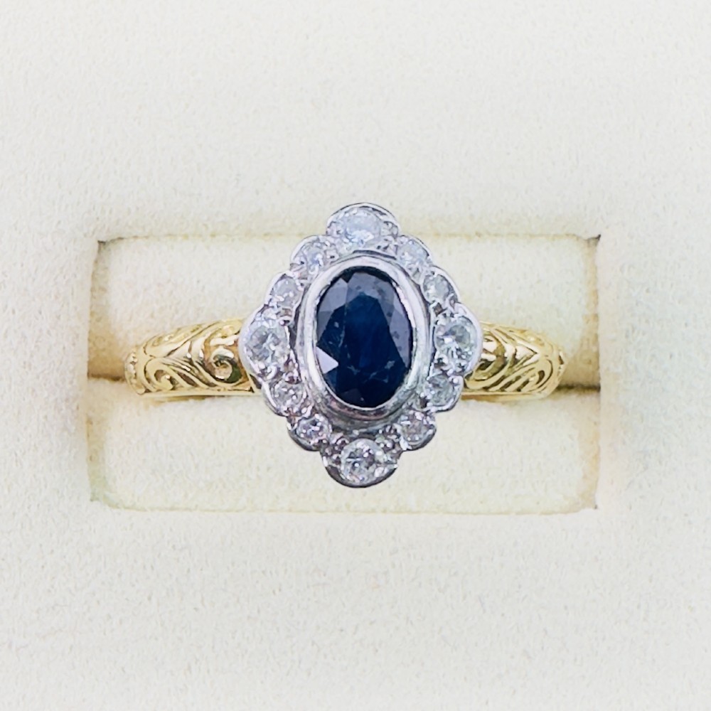 An 18ct yellow gold ring featuring a central oval blue sapphire surrounded by a border of small diamonds, set on a carved scrollwork band. View II