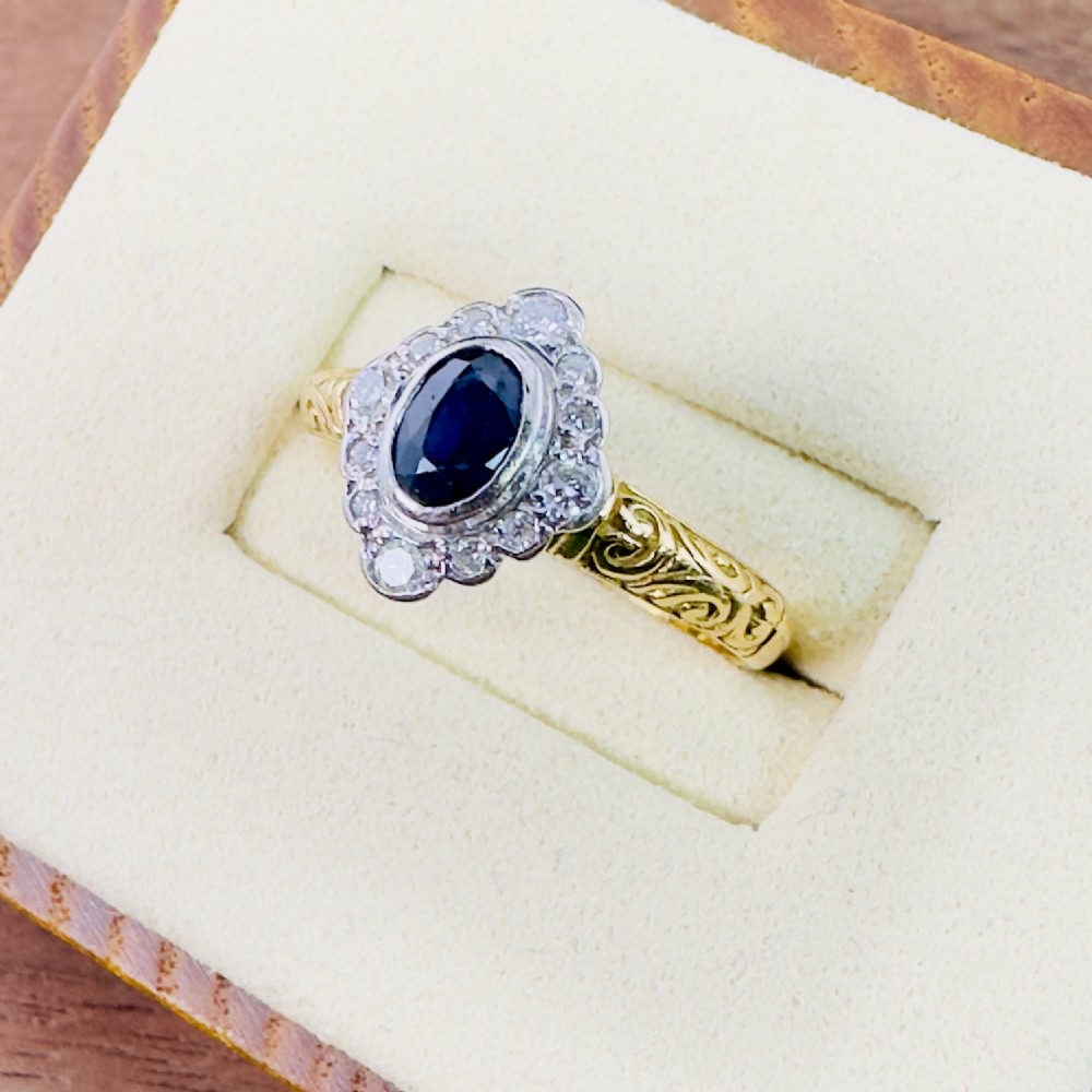 An 18ct yellow gold ring featuring a central oval blue sapphire surrounded by a border of small diamonds, set on a carved scrollwork band. View III
