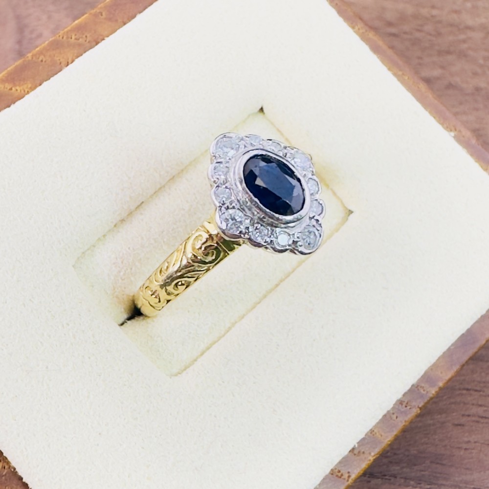 An 18ct yellow gold ring featuring a central oval blue sapphire surrounded by a border of small diamonds, set on a carved scrollwork band. View IV