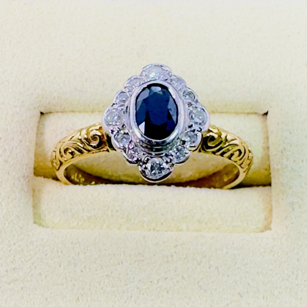 An 18ct yellow gold ring featuring a central oval blue sapphire surrounded by a border of small diamonds, set on a carved scrollwork band. View V