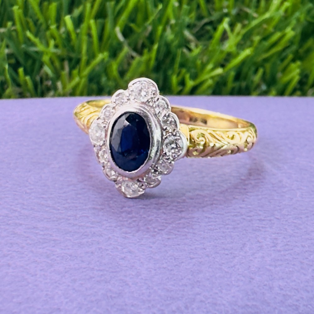 An 18ct yellow gold ring featuring a central oval blue sapphire surrounded by a border of small diamonds, set on a carved scrollwork band. View VII