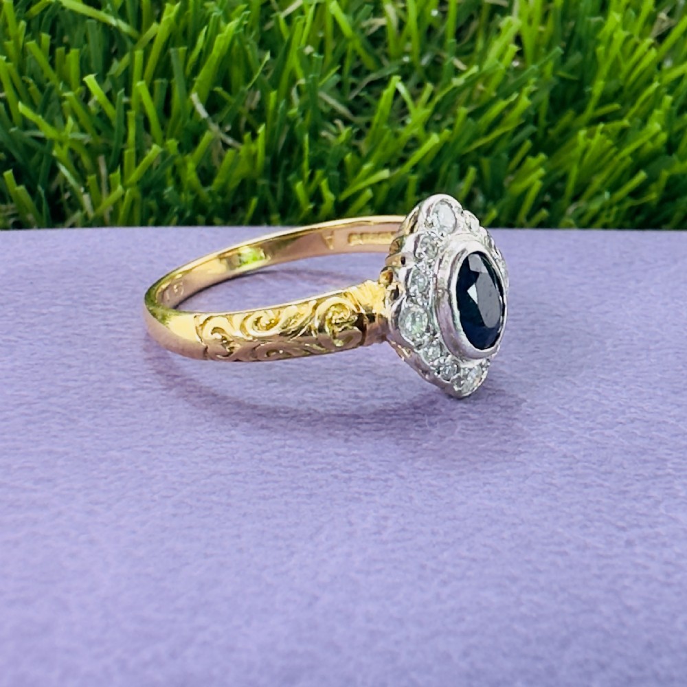 An 18ct yellow gold ring featuring a central oval blue sapphire surrounded by a border of small diamonds, set on a carved scrollwork band. View VIII
