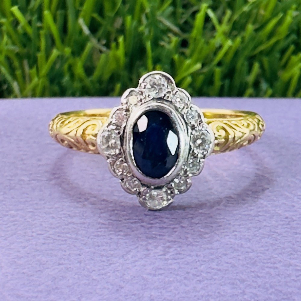 An 18ct yellow gold ring featuring a central oval blue sapphire surrounded by a border of small diamonds, set on a carved scrollwork band.
