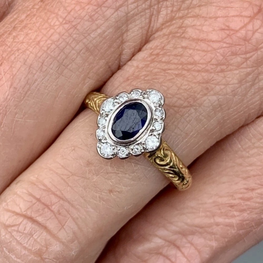 An 18ct yellow gold ring featuring a central oval blue sapphire surrounded by a border of small diamonds, set on a carved scrollwork band. View IX