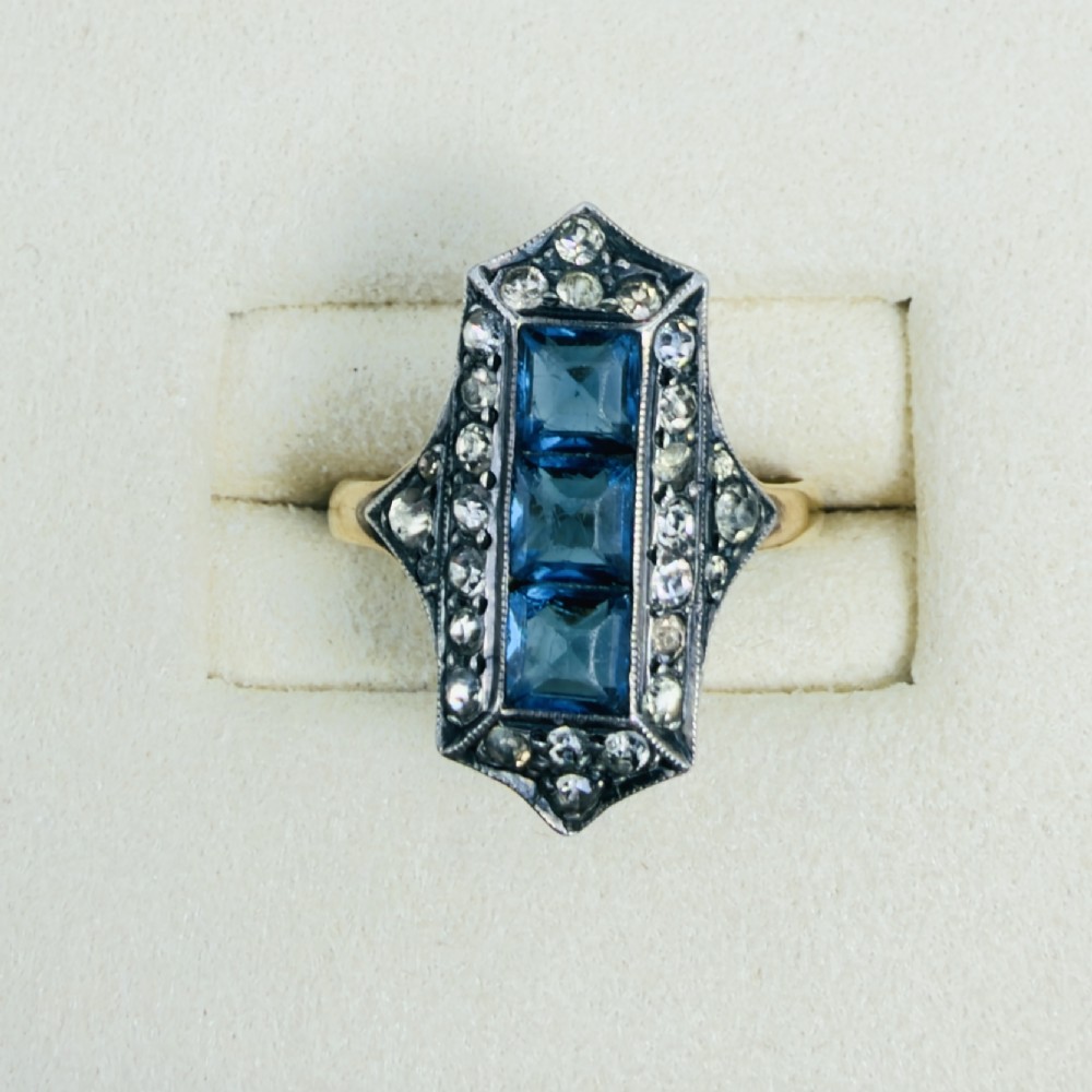 An Art Deco dress ring featuring an elongated geometric silver top set with three rectangular blue stones and bordered by round white paste stones on a yellow gold band. View II