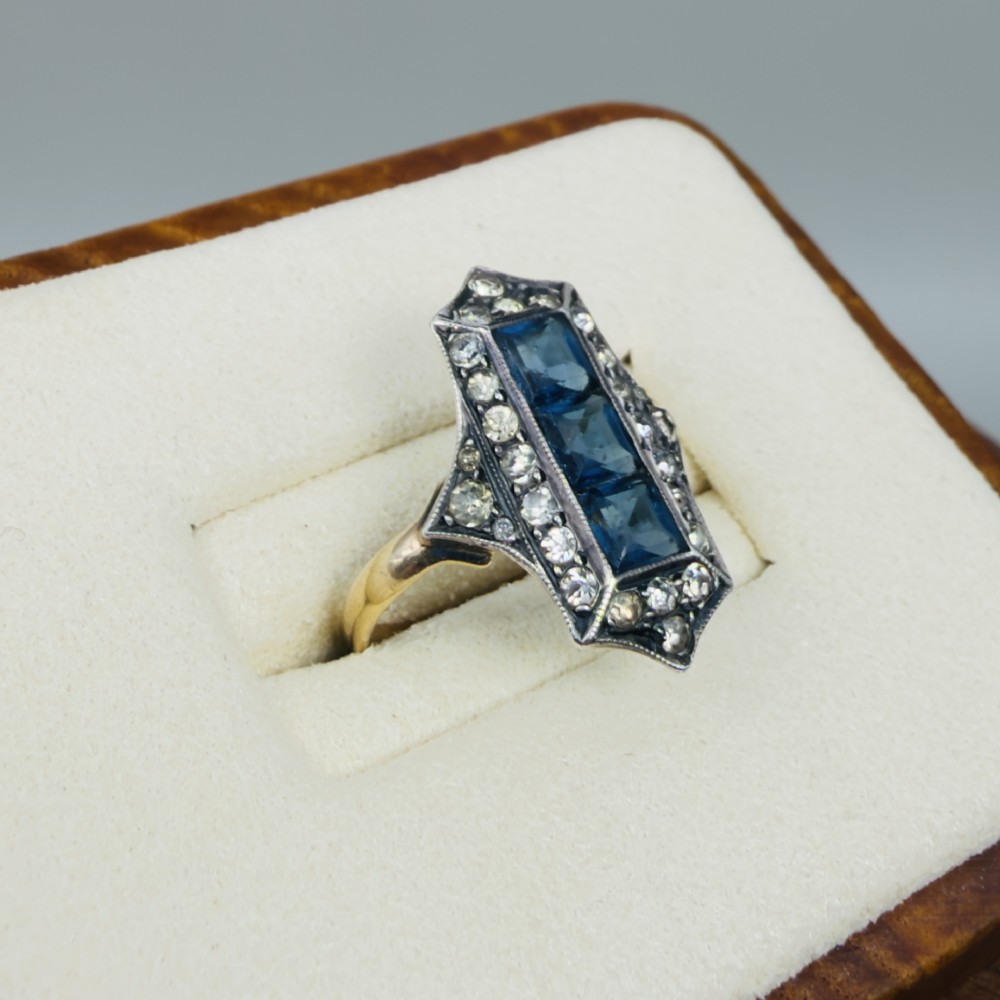An Art Deco dress ring featuring an elongated geometric silver top set with three rectangular blue stones and bordered by round white paste stones on a yellow gold band. View IV