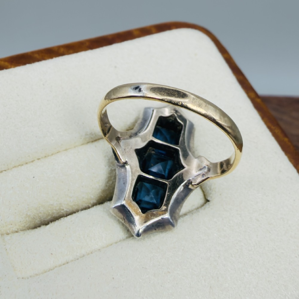 An Art Deco dress ring featuring an elongated geometric silver top set with three rectangular blue stones and bordered by round white paste stones on a yellow gold band. View V