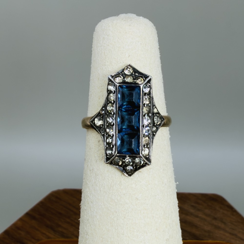 An Art Deco dress ring featuring an elongated geometric silver top set with three rectangular blue stones and bordered by round white paste stones on a yellow gold band. View VI