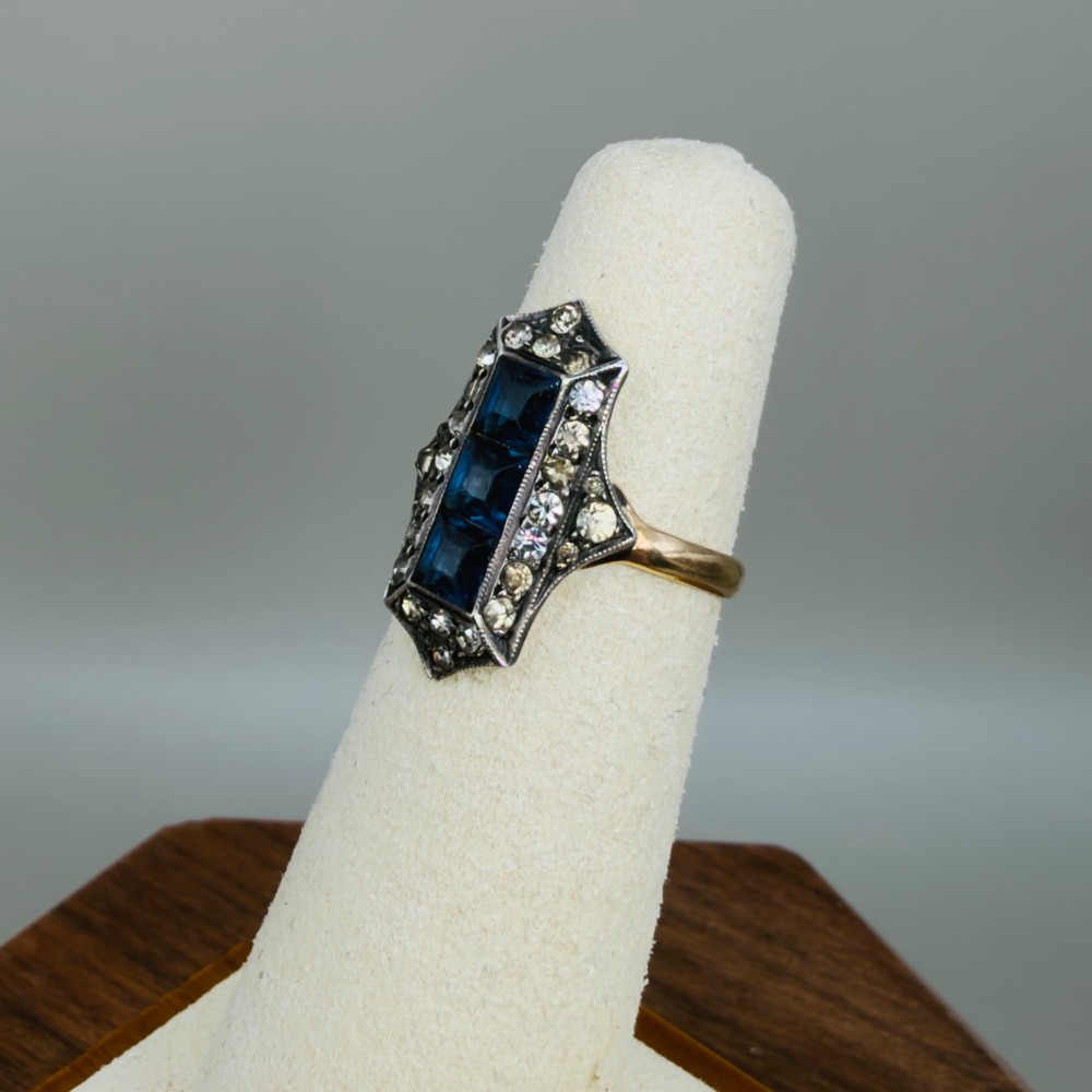An Art Deco dress ring featuring an elongated geometric silver top set with three rectangular blue stones and bordered by round white paste stones on a yellow gold band. View VII