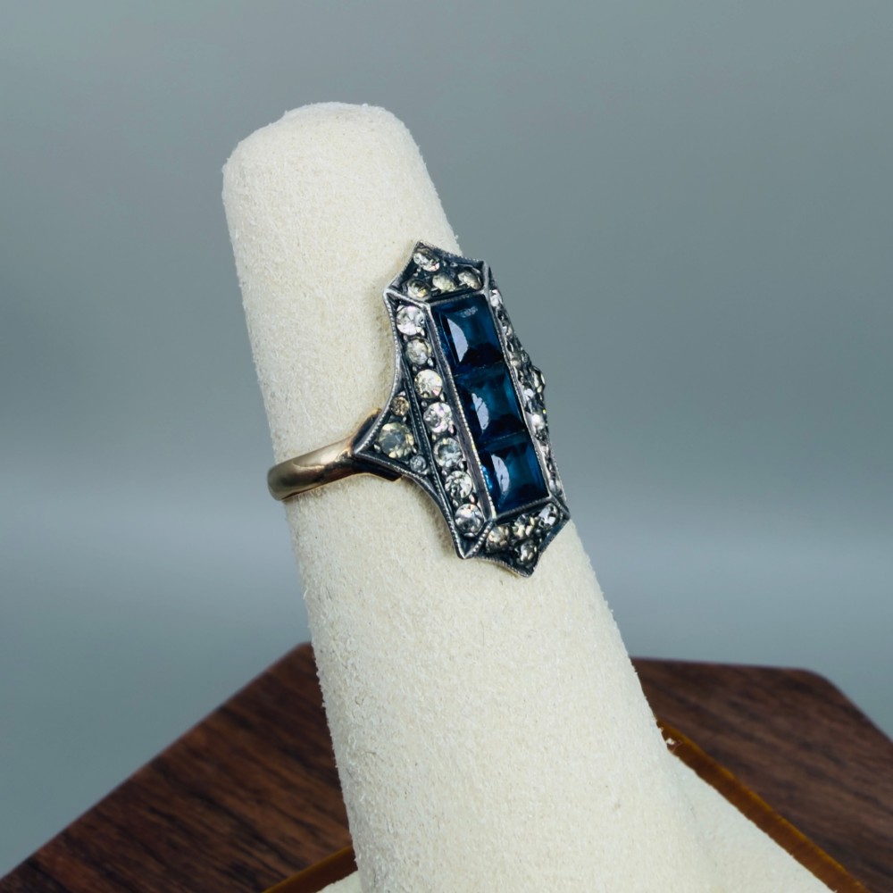 An Art Deco dress ring featuring an elongated geometric silver top set with three rectangular blue stones and bordered by round white paste stones on a yellow gold band. View VIII