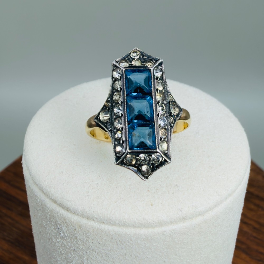 An Art Deco dress ring featuring an elongated geometric silver top set with three rectangular blue stones and bordered by round white paste stones on a yellow gold band.