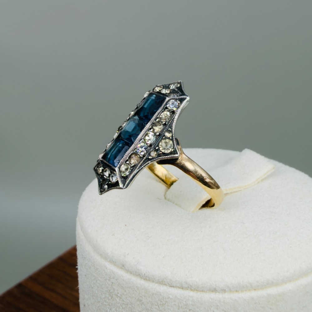 An Art Deco dress ring featuring an elongated geometric silver top set with three rectangular blue stones and bordered by round white paste stones on a yellow gold band. View IX