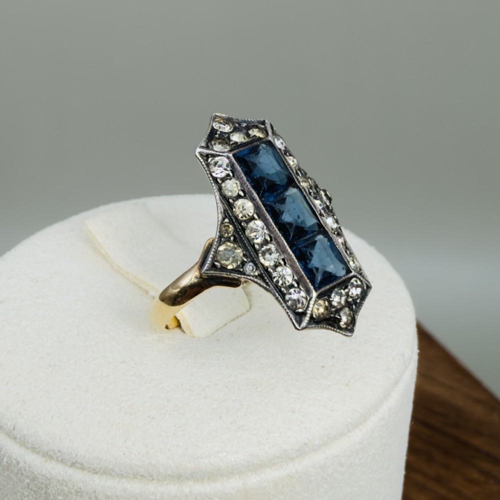 An Art Deco dress ring featuring an elongated geometric silver top set with three rectangular blue stones and bordered by round white paste stones on a yellow gold band. View X