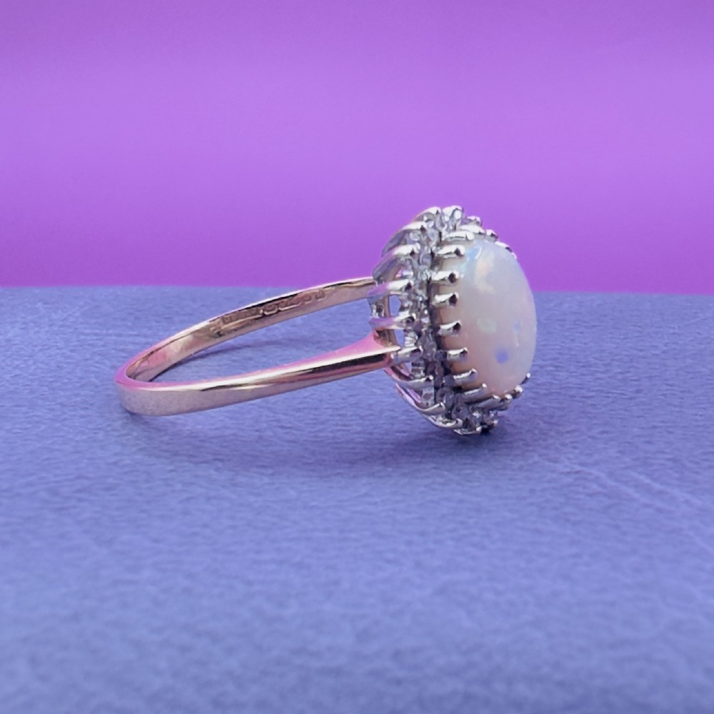 A 9ct yellow gold ring featuring a central oval cabochon opal surrounded by a halo of small claw-set diamonds. View X