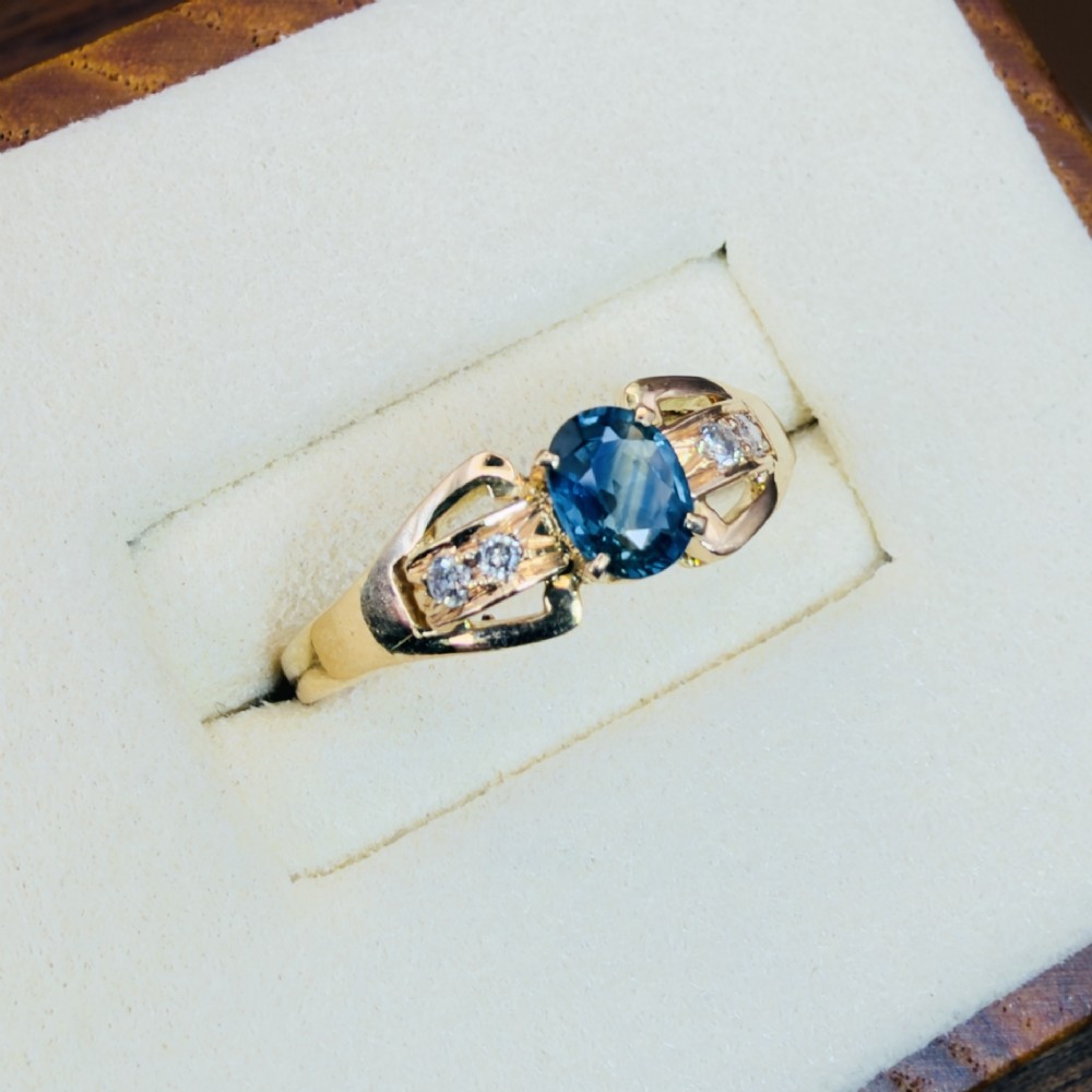 An 18ct yellow gold ring featuring a central oval-cut blue sapphire flanked by round diamonds set into carved shoulders. View IV