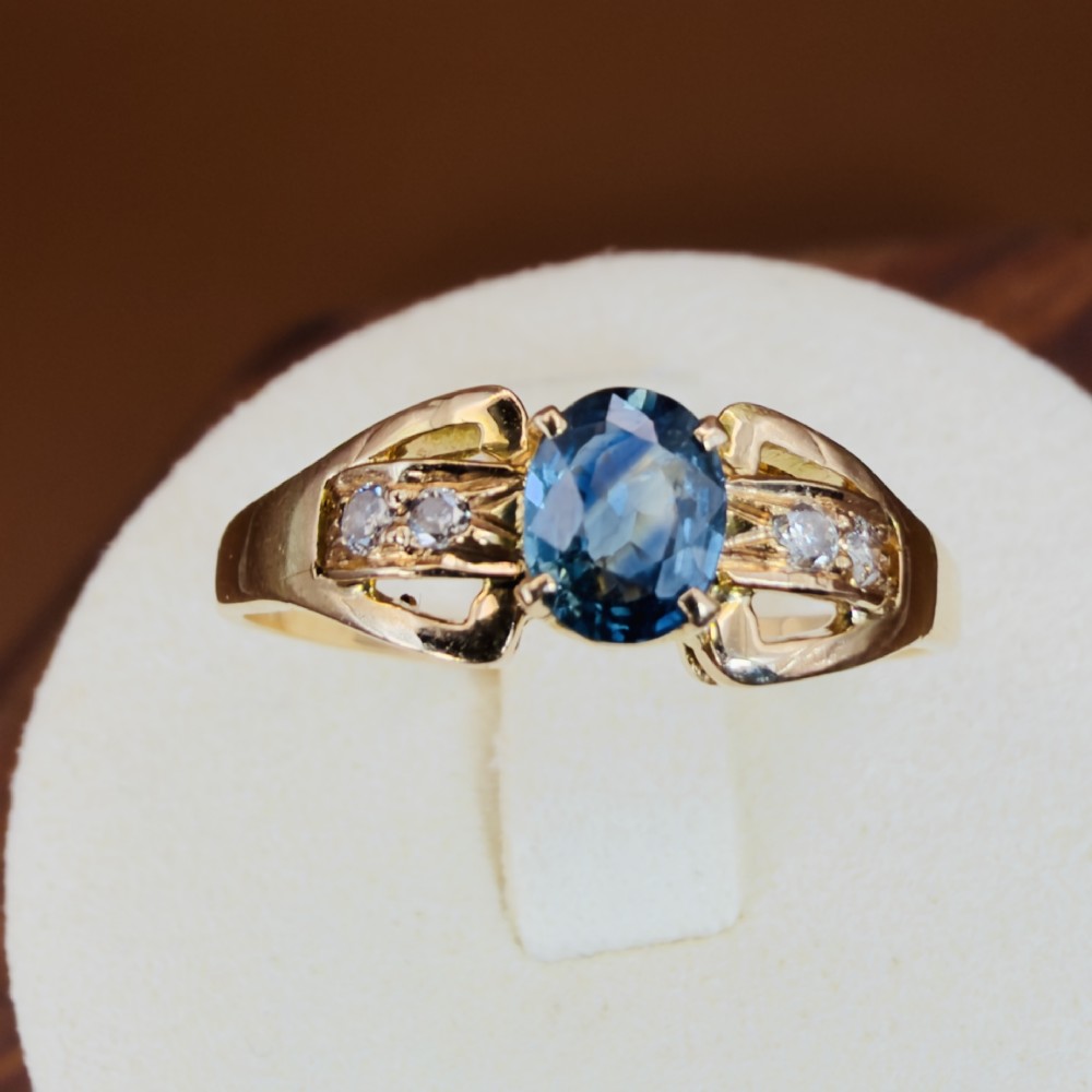 An 18ct yellow gold ring featuring a central oval-cut blue sapphire flanked by round diamonds set into carved shoulders.