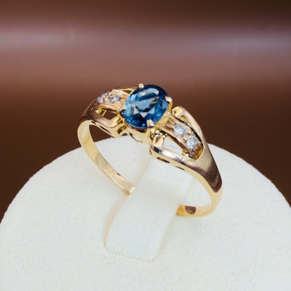 An 18ct yellow gold ring featuring a central oval-cut blue sapphire flanked by round diamonds set into carved shoulders. View IX