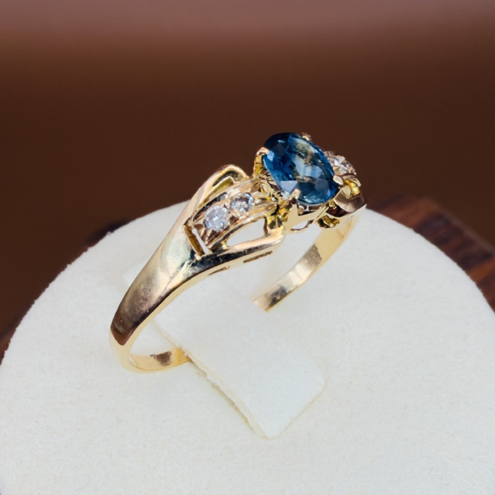 An 18ct yellow gold ring featuring a central oval-cut blue sapphire flanked by round diamonds set into carved shoulders. View X