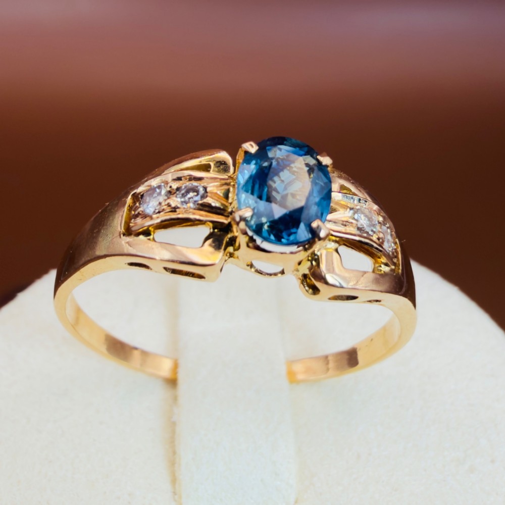 An 18ct yellow gold ring featuring a central oval-cut blue sapphire flanked by round diamonds set into carved shoulders. View XI