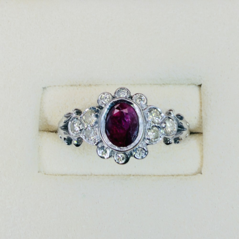 An 18ct white gold cluster ring featuring a central oval bezel-set red ruby framed by small round diamonds and decorative shoulders. View II