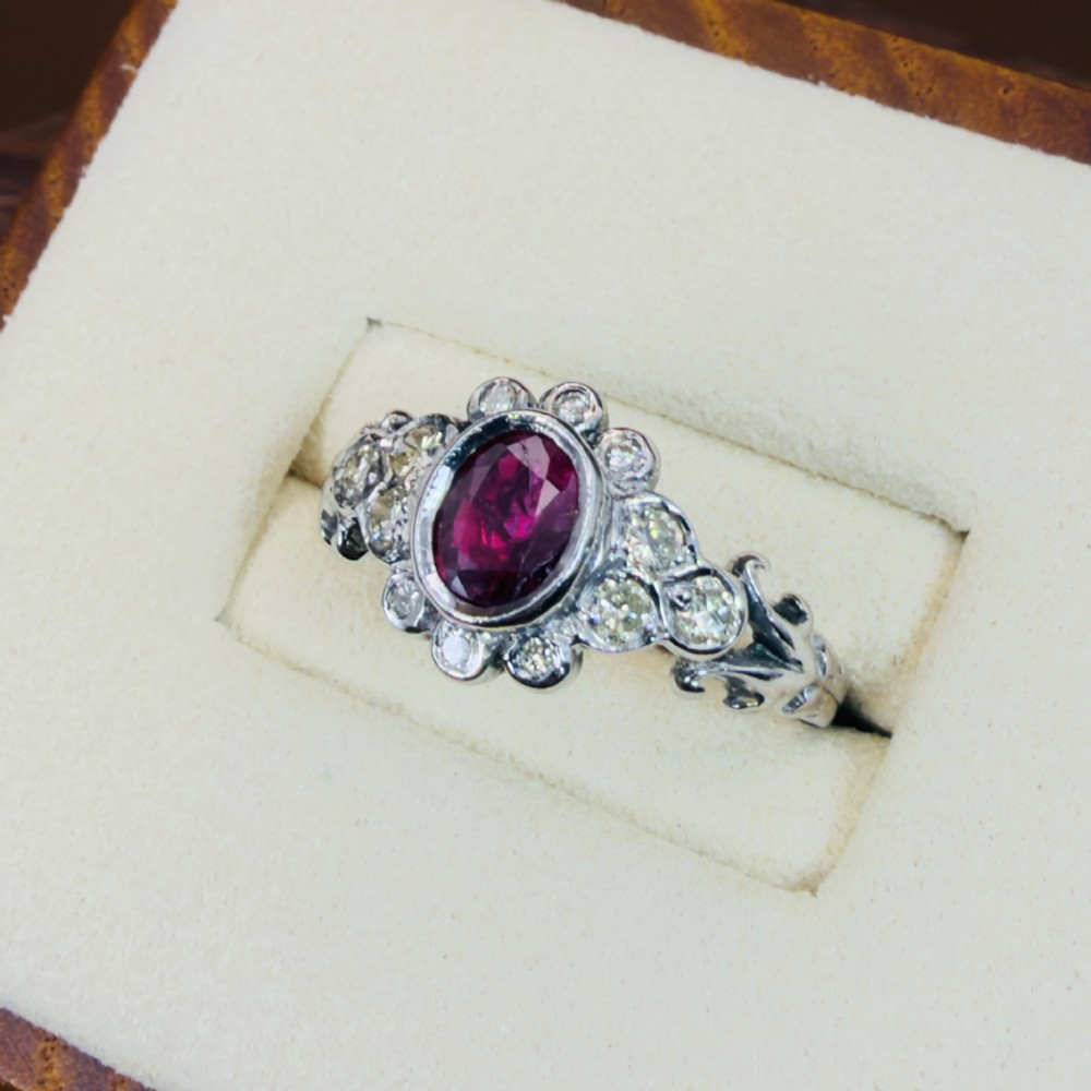 An 18ct white gold cluster ring featuring a central oval bezel-set red ruby framed by small round diamonds and decorative shoulders. View III