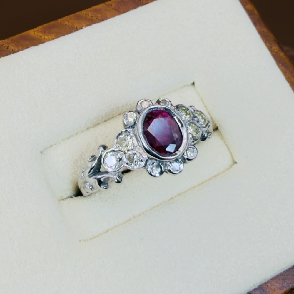 An 18ct white gold cluster ring featuring a central oval bezel-set red ruby framed by small round diamonds and decorative shoulders. View IV