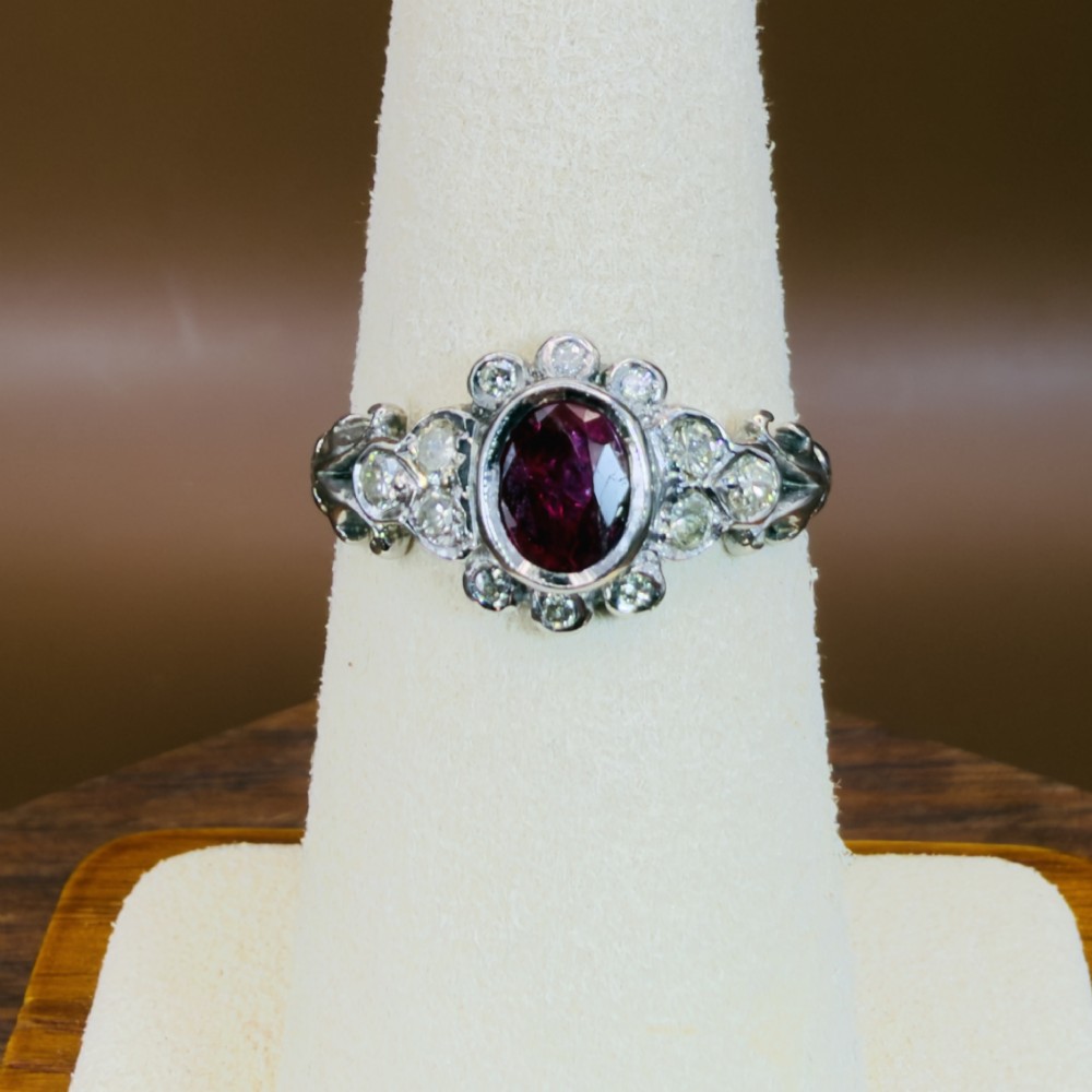 An 18ct white gold cluster ring featuring a central oval bezel-set red ruby framed by small round diamonds and decorative shoulders. View V