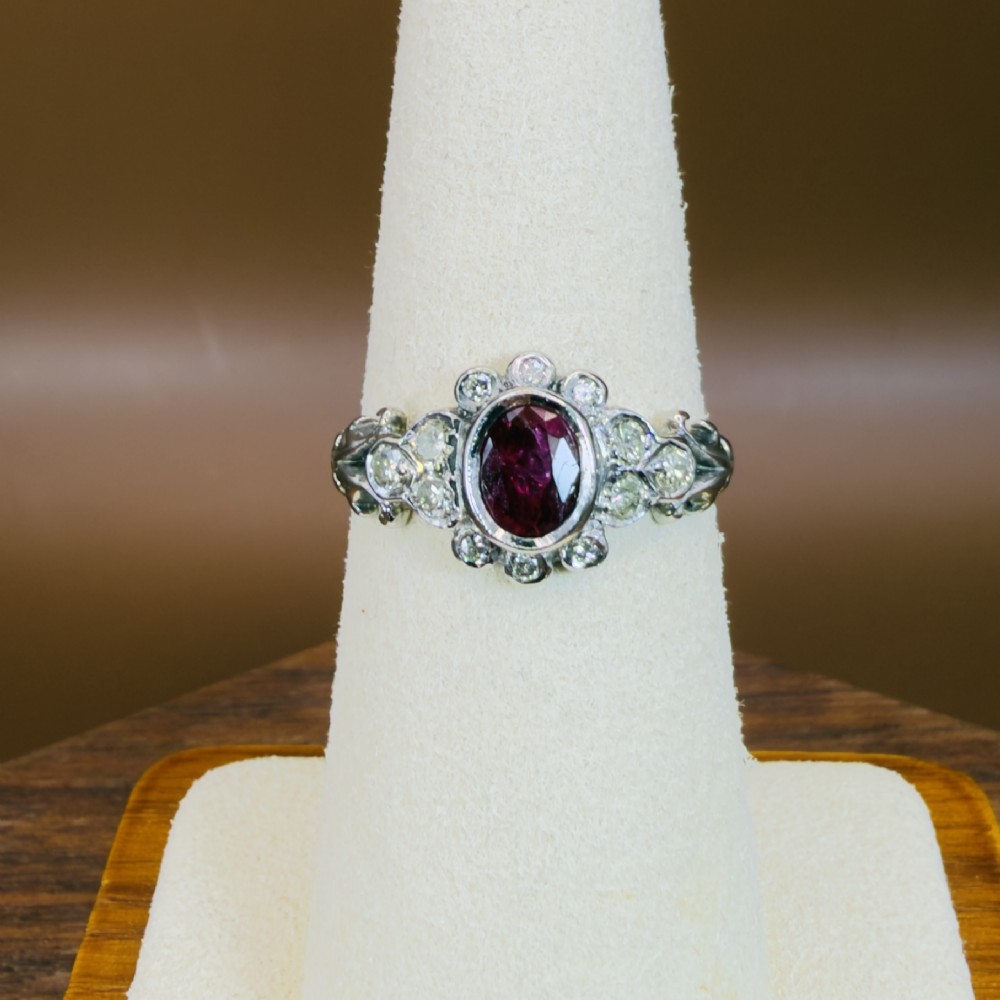 An 18ct white gold cluster ring featuring a central oval bezel-set red ruby framed by small round diamonds and decorative shoulders. View VI