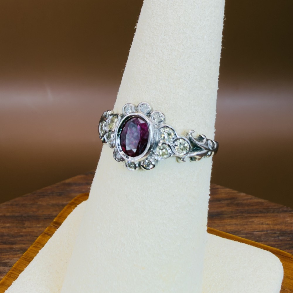 An 18ct white gold cluster ring featuring a central oval bezel-set red ruby framed by small round diamonds and decorative shoulders. View VII