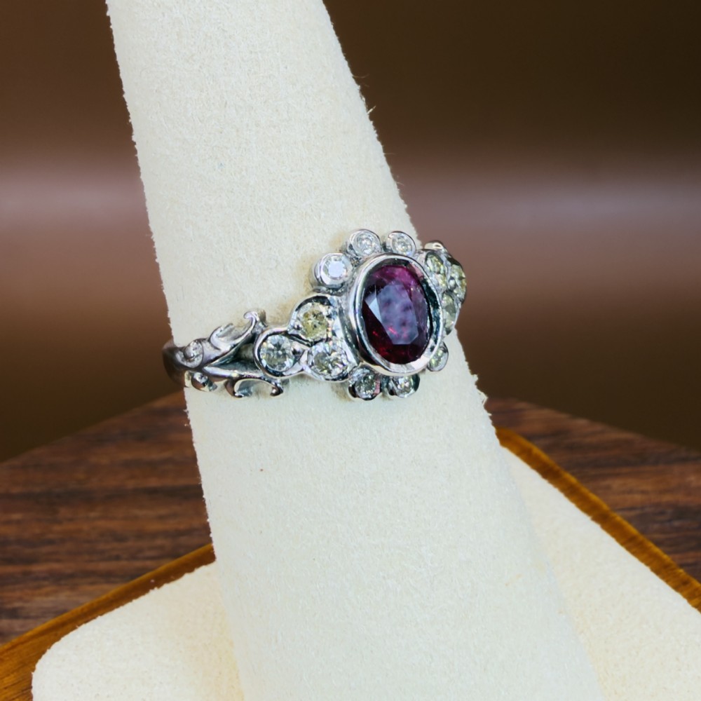 An 18ct white gold cluster ring featuring a central oval bezel-set red ruby framed by small round diamonds and decorative shoulders. View VIII